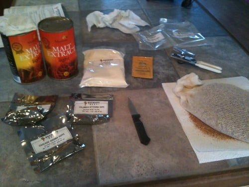 A kitchen counter with home brewing supplies including cans of malt extract, bags of hops, a packet of yeast, a knife, and a cloth bag with grains, alongside some cleaning tissues.