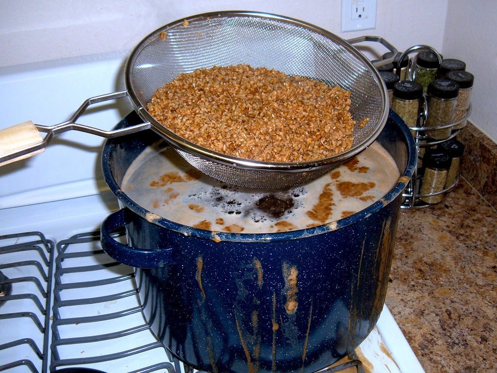 A mess on a kitchen stove where a blue pot has boiled over with what appears to be grains or oats, and a metal strainer above the pot is holding the same material.