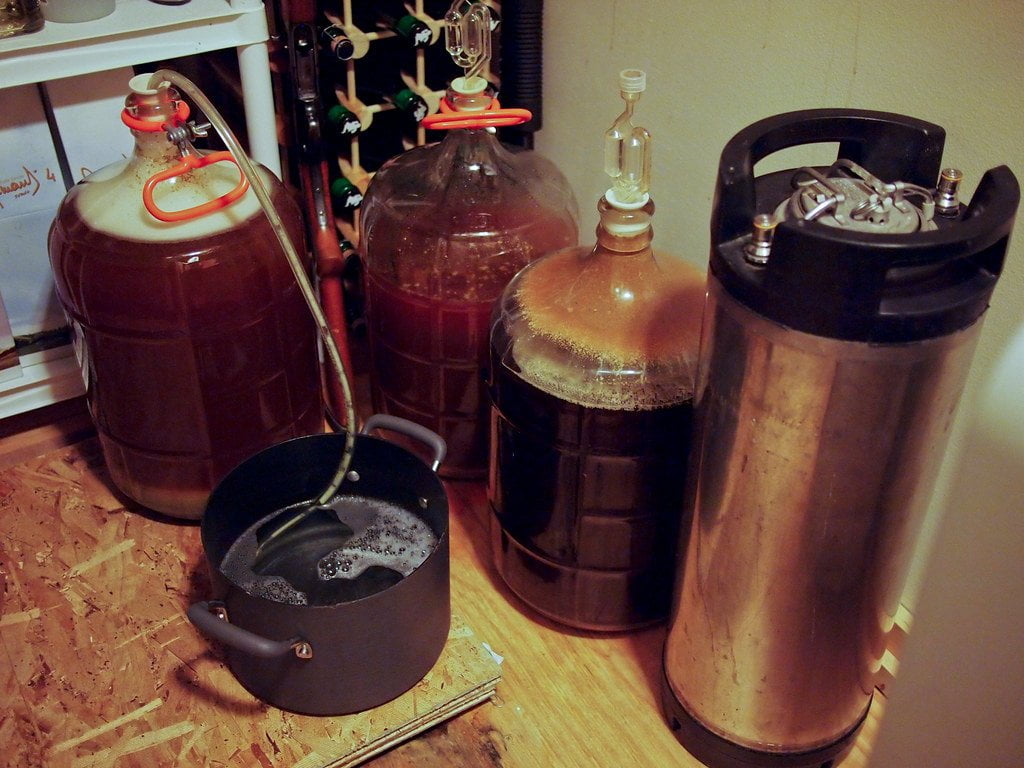 Homebrew beer equipment with three carboys fermenting beer and a stainless steel keg on a wooden floor next to a black pot filled with liquid.