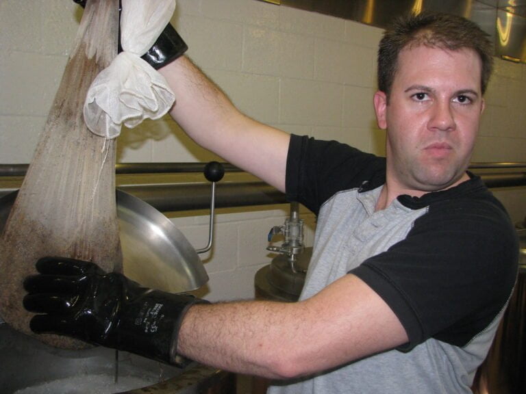 A man in a kitchen wearing black gloves is wringing out a cloth bag over a metal container.