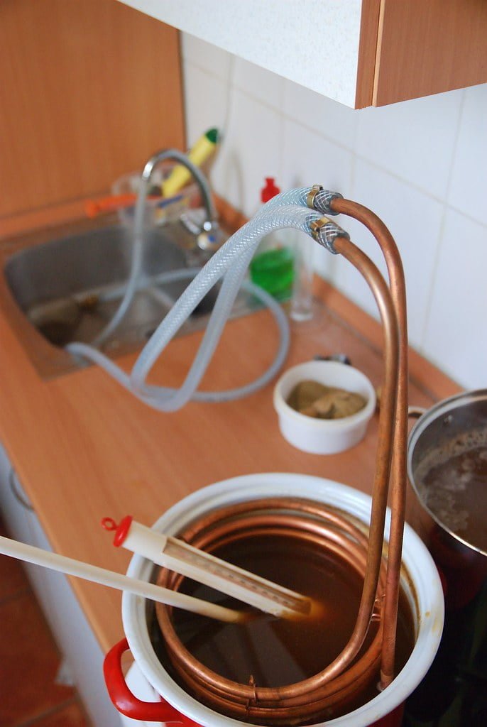 A homemade distillation setup on a kitchen counter with a copper coil emerging from a pot, passing through a water-filled sink for cooling, and leading into a collection vessel.