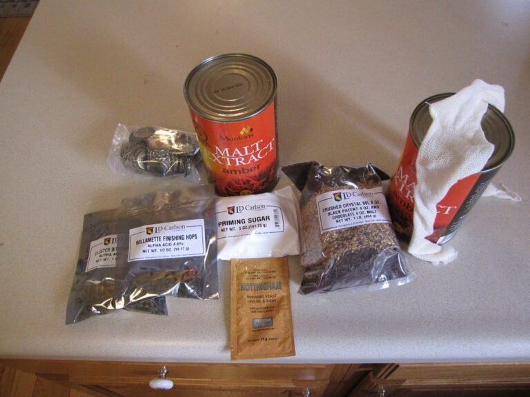 A kitchen countertop displaying various home brewing ingredients and supplies including malt extract canisters, bags of hops and grain, priming sugar, brewing yeast, and a cheesecloth bag.