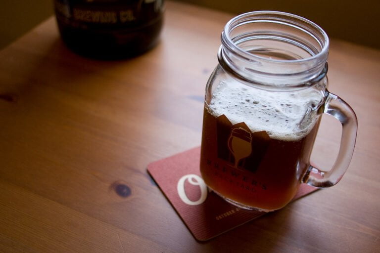A mason jar mug filled with amber-colored beer, resting on a coaster with the text "Brewer's Backyard" on a wooden table, with a blurred growler in the background.