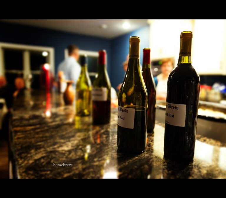 A focused view of several wine bottles with homemade labels on a marble countertop, with blurred figures of people in the background inside a kitchen.