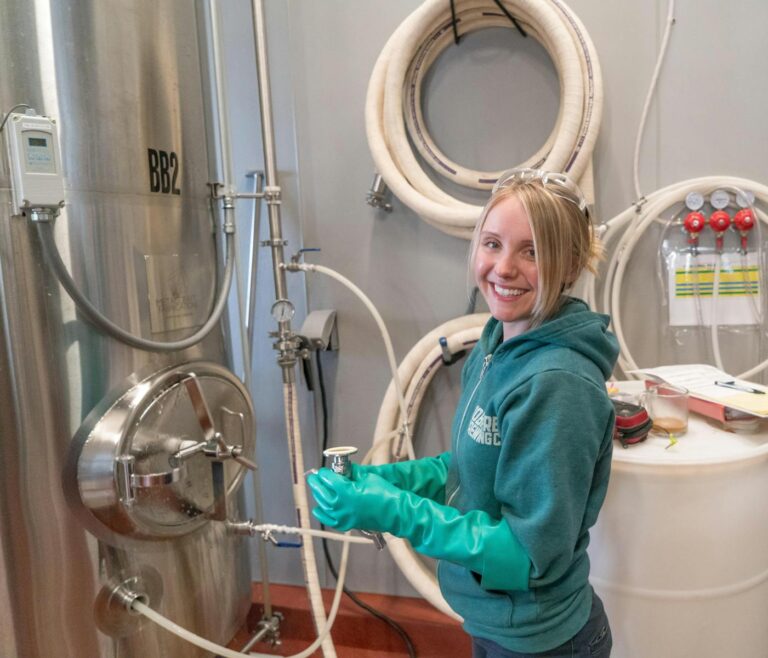 A smiling woman in a green hoodie and protective gloves working with equipment at a brewery.