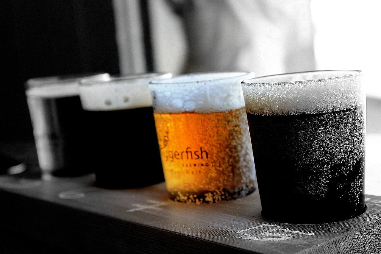 Four glasses of beer with varying colors and foam levels, displayed in a row on a wooden surface. The second glass is in focus, showing the label "Tigerfish Brewing." Other glasses are out of focus. The background is desaturated, highlighting the amber color of the beer in the focused glass.