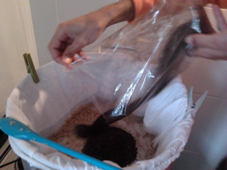A person is pouring coffee grounds from a plastic bag into a white fabric-lined container, with a wooden spoon visible in the foreground and a green clothespin attached to the container.
