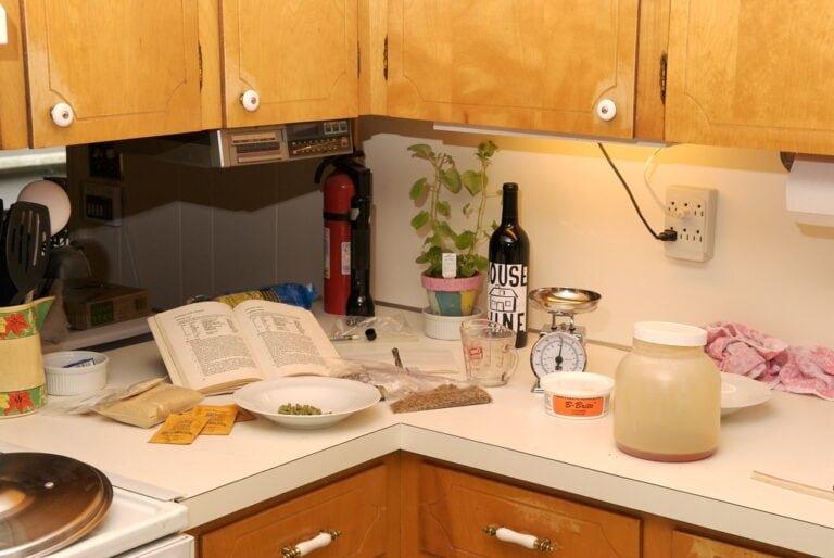 A cluttered kitchen countertop with an open cookbook, ingredients like peas and spices, cooking utensils, a bottle labeled "HOUSE WINE," a scale, a measuring cup, and a large jar, possibly containing a fermenting substance, under wooden cabinets. There is an unplugged power strip and a fire extinguisher in the background.