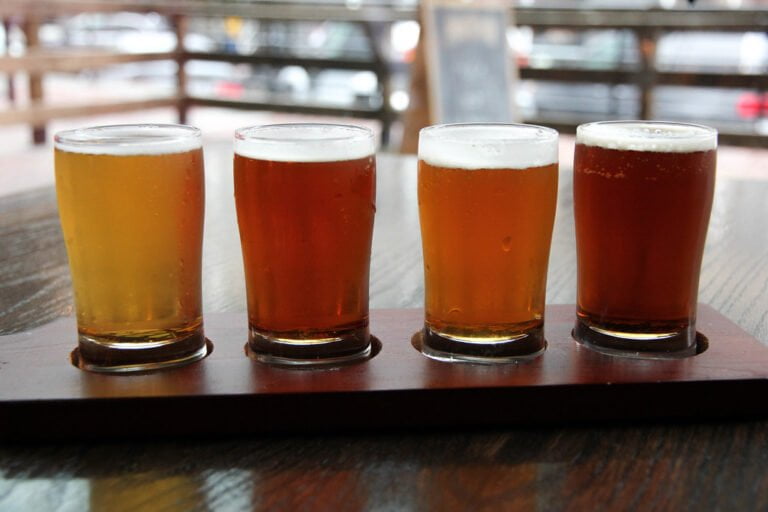 Four glasses of beer with varying colors and froth levels, presented on a wooden flight paddle on a table.
