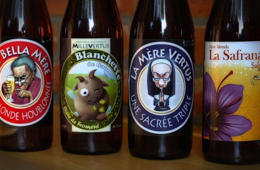 Four bottles of beer with illustrated labels displayed side by side. From left to right, the labels feature a cartoon of an elderly woman drinking beer, a smiling pig holding hops, an angry elderly nun, and a large purple flower, respectively. Each label also contains text in French describing the beer type and brand.