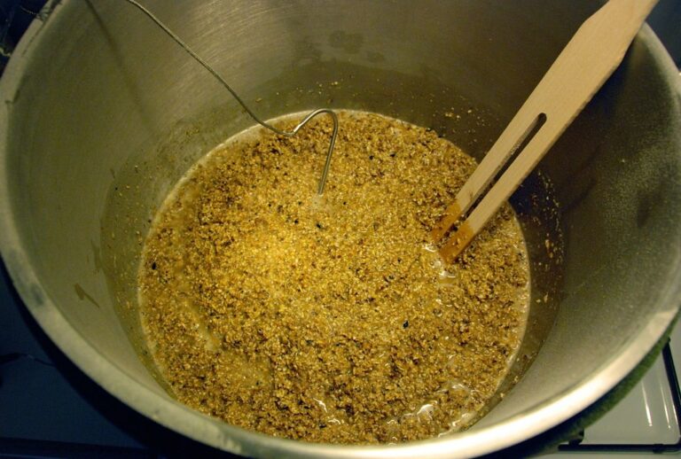 A large metal brewing kettle containing a mixture of grains and liquids during the mashing process of beer brewing, with a wooden spoon and a stainless steel stirring device.