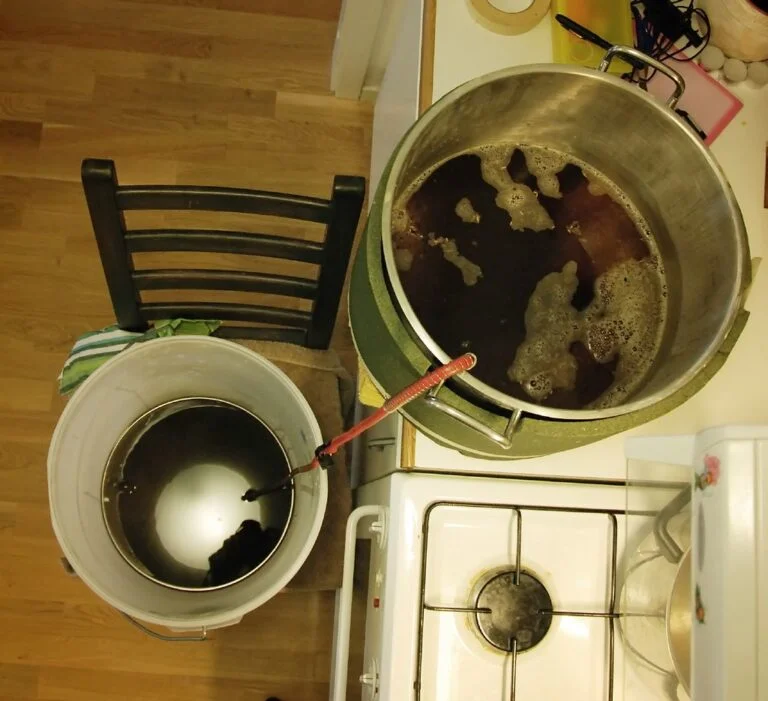 A top-down view of a kitchen with a large pot on the stove containing a dark liquid with foam on top, next to it is a bucket with similar liquid connected by a red hose to the pot, and a wooden chair nearby.
