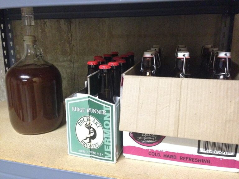 A glass carboy filled with a dark liquid and a six-pack of bottled beer next to a cardboard beer case on a metal storage shelf.