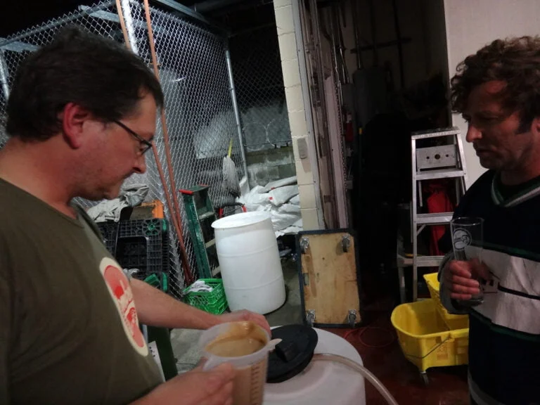 Two men standing in a cluttered storeroom, one holding a pint glass and the other examining a container with a creamy substance.