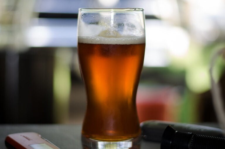 A close-up photo of a glass of amber beer with foam on top, resting on a table with a blurred background, with a cell phone and another object nearby.