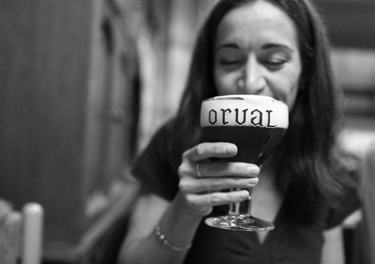 A woman smiling and holding a chalice glass of beer with "Orval" written on the foam, in a blurred black and white background.