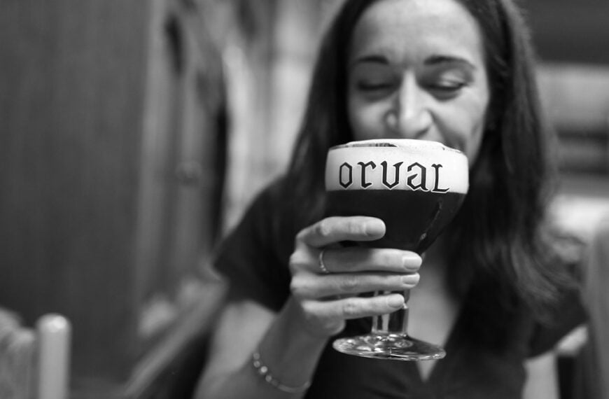 A woman smiling and holding a chalice glass of beer with "Orval" written on the foam, in a blurred black and white background.