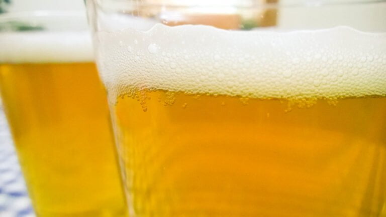 Close-up of two glasses of beer with a focus on the frothy heads, set against a blurry background.