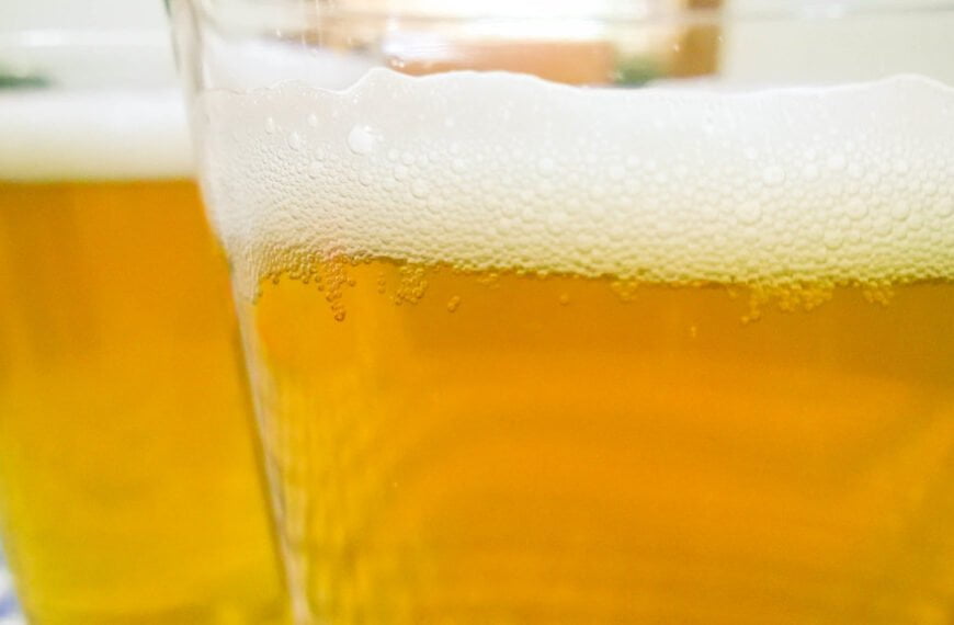 Close-up of two glasses of beer with a focus on the frothy heads, set against a blurry background.