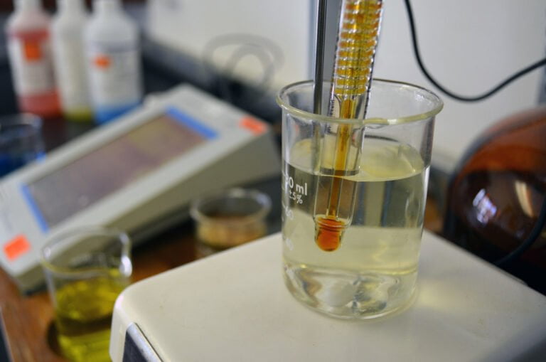 A hydrometer is submerged in a beaker of liquid on a digital scale, with lab bottles in the soft-focused background.