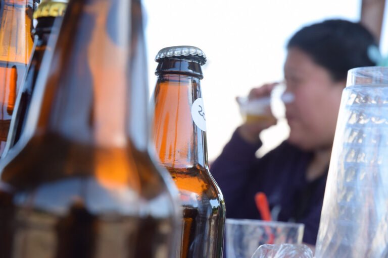 A blurred person is seen in the background drinking from a glass, while in focus in the foreground are empty beer bottles and a glass on a table.