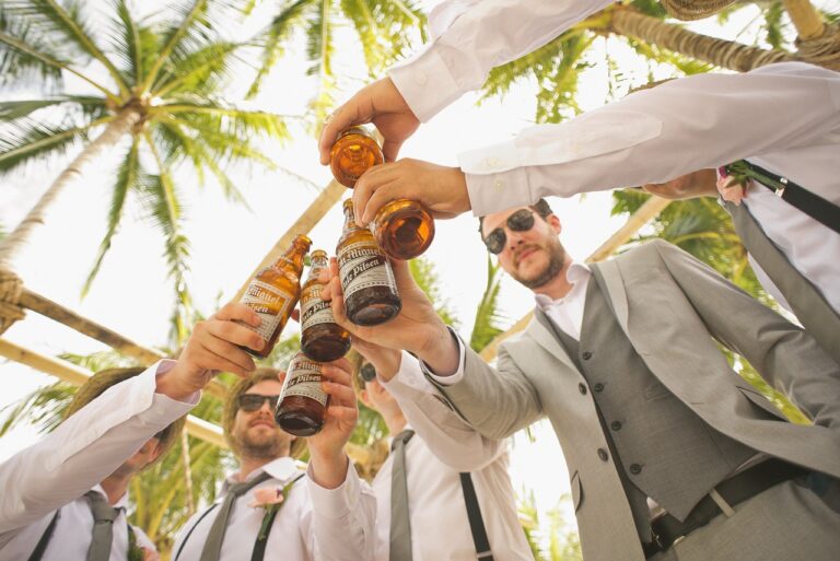 A group of men in formal attire toasting with beer bottles under palm trees.