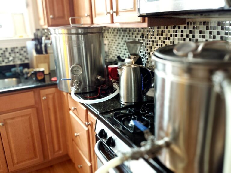 A homebrewing setup with stainless steel equipment on a kitchen stove, including a large brewing kettle with a spigot and attached tubing, and a smaller container, with a blurred background showing a tiled backsplash and wooden cabinets.