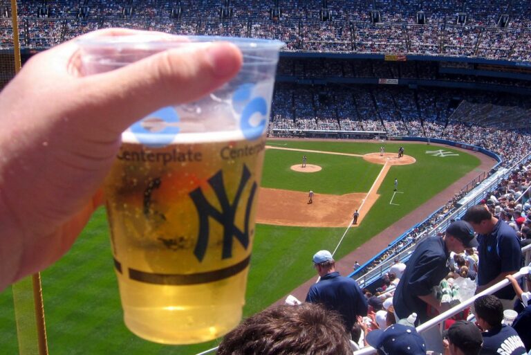A person holds a plastic cup of beer with the New York Yankees logo at a baseball stadium, with a view of the field and spectators in the stands during a sunny day.
