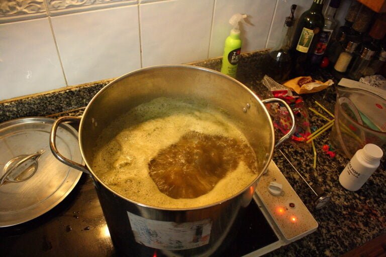 A large pot boiling on a kitchen stove with foam forming on top, next to various kitchen items and ingredients scattered on the counter.