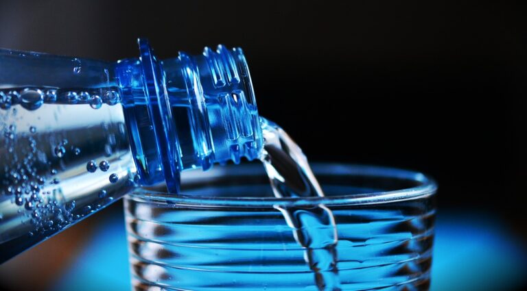 Clear water being poured from a plastic bottle into a glass cup, with a focus on the stream of liquid and the mouth of the bottle.