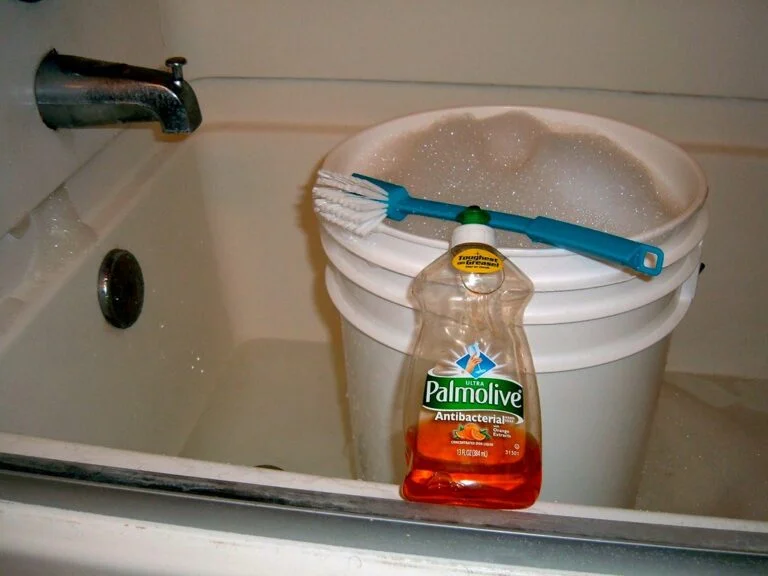 A white plastic bucket filled with soapy water and a scrub brush on top, next to a Palmolive antibacterial dish soap bottle, placed in a bathtub with the faucet slightly visible above.