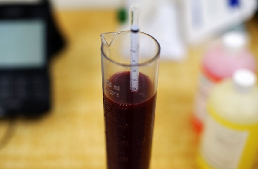 A pipette is inserted into a cylindrical glass container filled with a red liquid on a laboratory bench, with a blurred background showing lab equipment and bottles.