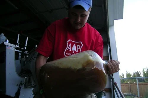 A person in a red t-shirt pouring a large container of liquid into a machine, indoors with a shelter-like structure in the background.