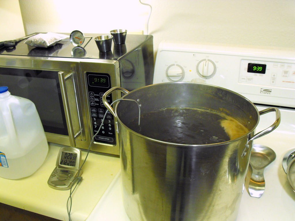 A kitchen countertop with a large stainless steel pot on a white stove. The pot appears to contain a liquid, possibly water. A digital thermometer, measuring cups, a gallon of milk, and a microwave displaying the time 9:39 are also on the countertop. There's a digital clock on the stove also showing the time 9:39.