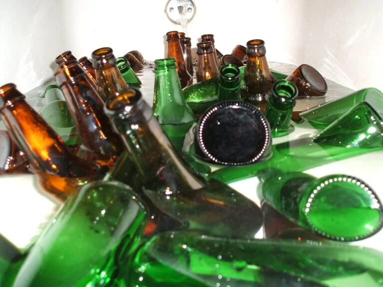 A collection of empty glass bottles in green and brown scattered haphazardly, some upright and others lying on their sides, with reflections visible on a shiny surface.