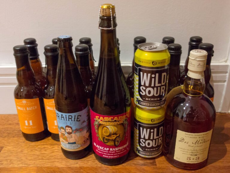 A variety of alcoholic beverages including craft beers and a bottle of aged rum on a table. The beers feature colorful labels from brands like Prairie Artisan Ales and Cascade Brewing, with flavors like tropical sour and blackcap raspberry.