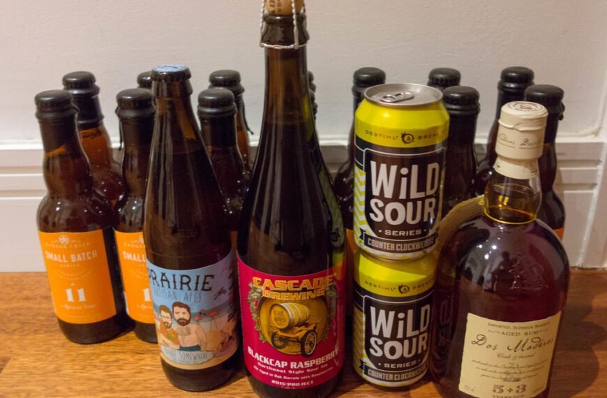 A variety of alcoholic beverages including craft beers and a bottle of aged rum on a table. The beers feature colorful labels from brands like Prairie Artisan Ales and Cascade Brewing, with flavors like tropical sour and blackcap raspberry.