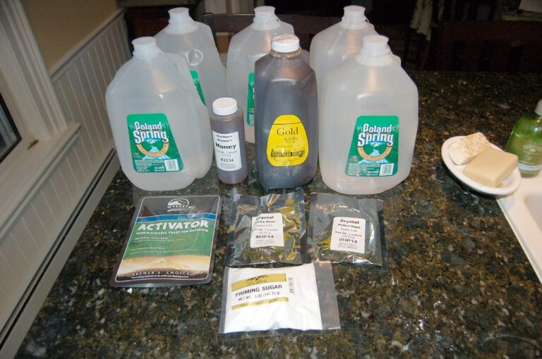 Several items on a kitchen countertop, including three gallon jugs of Poland Spring water, two containers labelled "Gold" and "Northern Honey," bags labelled "ACTIVATOR" yeast, "Crystal" hops, and "PRIMING SUGAR," as well as two bars of butter and a dish with two wedges of cheese.