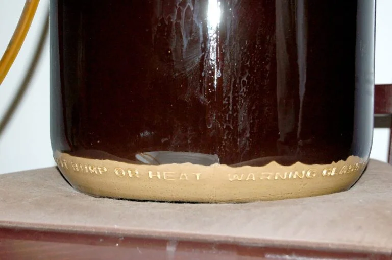 A close-up of a dark liquid inside a glass carboy with a warning label embossed at the bottom reading "NO BUMP OR HEAT WARNING GLASS," placed on a brown surface with a light brown sediment layer at the base of the liquid. A tube is visible exiting the top of the carboy.