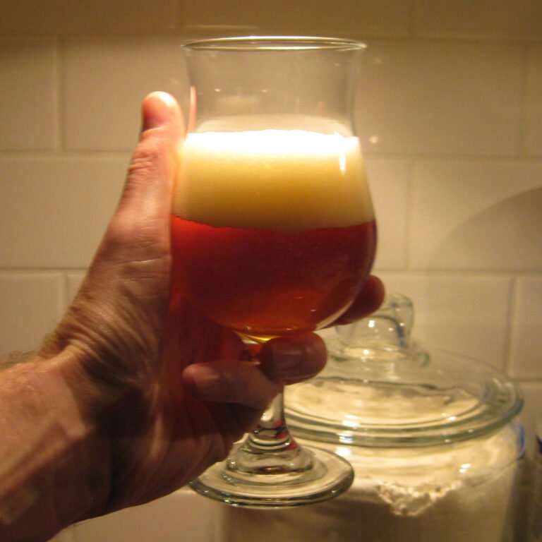 A person's hand holding a tulip-shaped glass of amber beer with a thick white foam head, in front of a tiled kitchen backsplash.