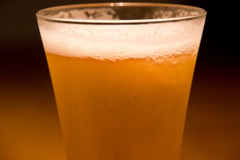 A close-up of a glass of beer with a frothy head, set against a dark background.
