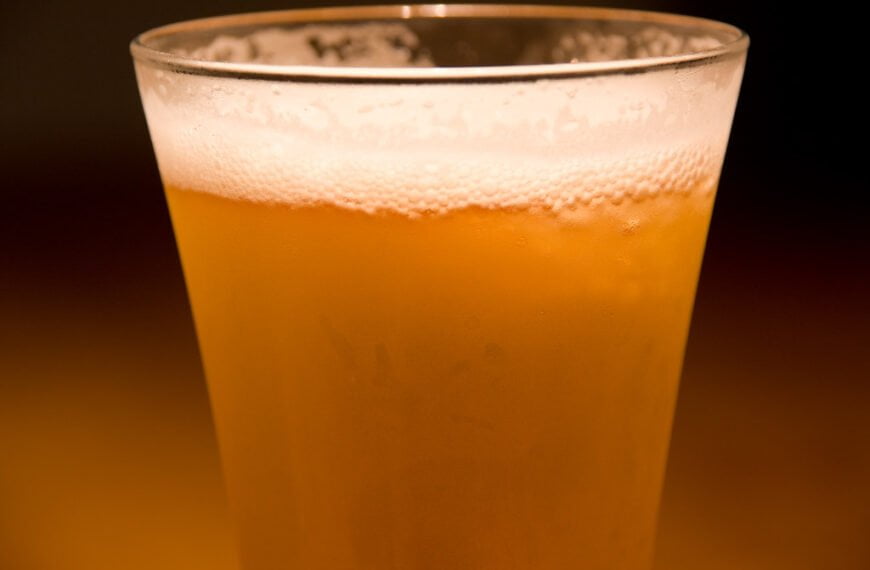 A close-up of a glass of beer with a frothy head, set against a dark background.