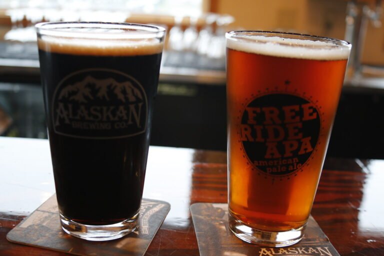 Two glasses of beer on coasters, one dark stout with "Alaskan Brewing Co." logo and one amber ale with "Free Ride APA American pale ale" text, on a wooden surface with a blurred background.