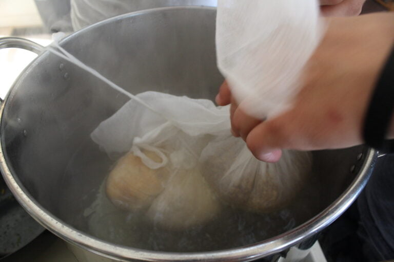 A person's hand is lifting a cheesecloth bag containing ingredients out of a steamy pot.