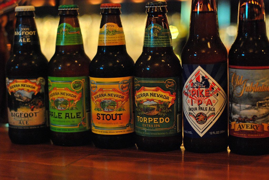 A row of assorted craft beer bottles on a wooden surface, with labels facing forward showing different types of ale, including Sierra Nevada Bigfoot Ale, Pale Ale, Stout, Torpedo Extra IPA, Pike IPA, and Avery Old Jubilation.