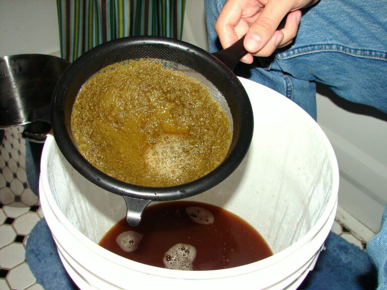 Person straining a brown liquid through a mesh strainer into a white bucket.