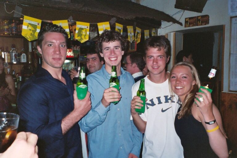Four young adults smiling and holding beer bottles in a crowded bar with festive decorations.