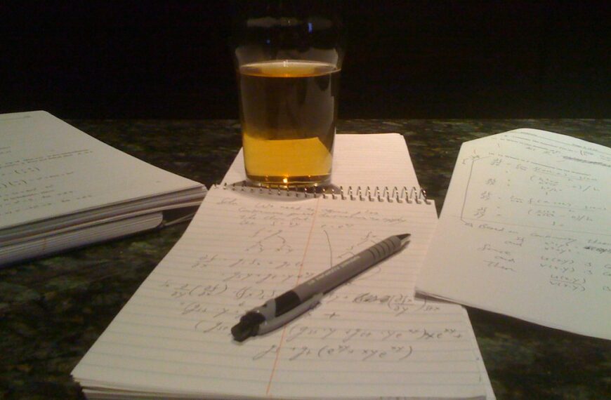 A glass of beer on a table with scattered papers containing mathematical equations and a pen.