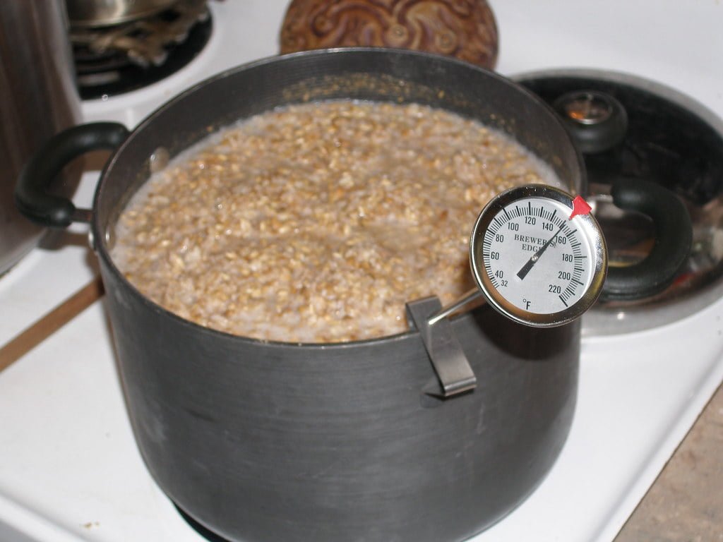 A pot filled with oatmeal is being cooked on a stove with a thermometer inserted showing a temperature around 140 degrees Fahrenheit.