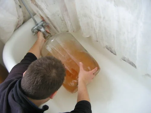 A person filling a large glass jar with amber-colored liquid from a bathtub faucet.